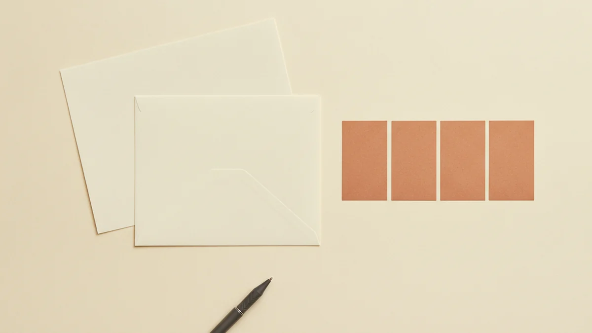 A blank cream letter and envelope on a cream desk beside a row of plain terracotta paper squares and a charcoal pen