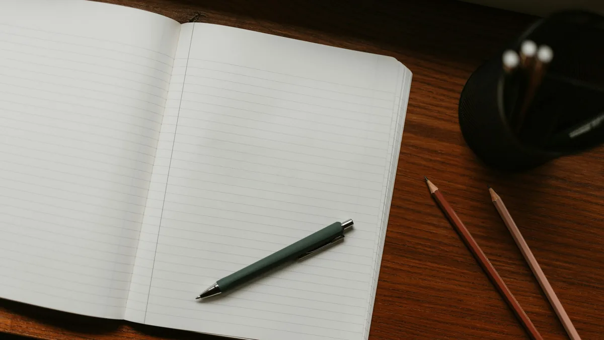 An open blank ruled notebook with a pen resting on it, beside two pencils on a dark wooden desk