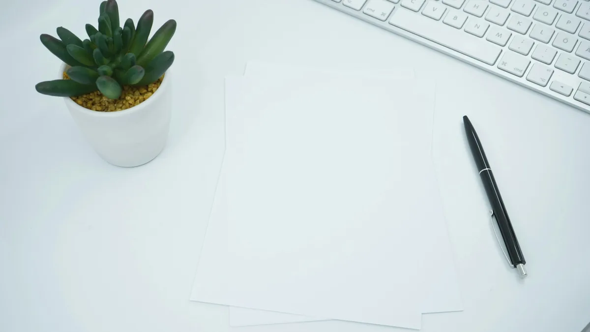Two blank sheets of paper on a white desk beside a pen, a keyboard and a small potted plant